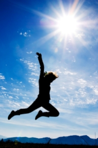 Person jumping with the sun, mountains, and a clear, blue sky in the background.